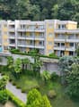 A large yellow apartment building with several balconies. Visible swimming pool. Trees, bushes, and greenery surround the building. In the distance a hilly background is visible.