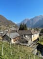 The image shows a picturesque mountain village nestled in a valley, with snow-capped peaks in the background. The village consists of traditional houses with red-tiled roofs, surrounded by vineyards and orchards. There are several trees and a grassy area