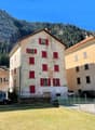 4 story house, red shutters, beige and stone wall, small garden, surrounded by green mountains