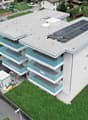 3-story apartment building, solar panels on roof, blue balconies, surrounding greenery, parking area