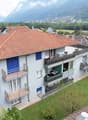 3-story building, red tiled roof, brown facade, balconies, satellite dish, surrounded by greenery, distant mountains, neighboring buildings with solar panels
