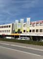 Office building with several floors, brick exterior, glass windows, multiple banners, cars parked in front, surrounding greenery, mountains in the background.