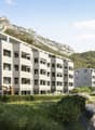 3-story building, white walls, large windows, balconies, green roof, garden, parking spots, grassy hills, mountains in the background