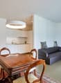 Open-concept living space with a dining area, kitchen, and living room. The dining table is made of wood with ornate chairs. The kitchen has white cabinets and a modern appliance, likely a dishwasher. The living room has a gray sofa and a decorative wall