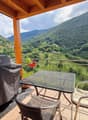 Furnished balcony with chairs, table, grill, plant, tiled floor, mesh railings, view of mountains and valley, hanging potted plant.