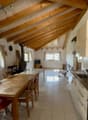 open dining area with wooden dining table, chairs, wooden beams on the ceiling, tiled floor, open kitchen with sink, appliances, small window with curtains, wooden cabinet, table and chair, black object, clock, and speaker.