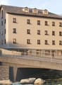 4-story building, beige facade, numerous windows with balconies, stone bridge over a river