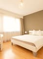 Spacious bedroom with hardwood flooring, beige walls, large windows with sheer curtains, a king-size bed, a white armchair, and a crystal chandelier overhead.