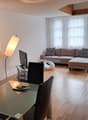 Spacious living room with large windows, hardwood floors, a brown sectional sofa with patterned throw pillows, a glass coffee table, and a floor lamp. The room has an open and airy feel.