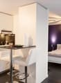 The image shows a studio apartment with an open-concept layout. The kitchen area features white cabinets, a black countertop, and a dishwasher. The living and sleeping areas are separated by a partial wall, with a bed visible in the bedroom section. The r