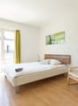 Spacious bedroom with large windows, wooden floor, white walls, a bed with wooden headboard, a comfortable armchair, and a colorful artwork on the wall.