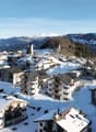 Mountain village covered in snow, several residential buildings, snowy mountains in the background, clear blue sky