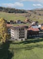 A large residential building with a modern architectural design, situated in a lush, green valley. The building has solar panels installed on its roof, and there are several houses nearby, some of which have attached garages. The surroundings are scenic,