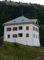 white house, red shutters, gray roof, on a hill, surrounded by trees