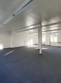 Large open office space with high ceilings, white walls, and gray carpeted flooring. The room has several support columns and large windows providing natural light. The space appears to be ready for use as an office or commercial space.