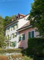 Two story house, white and pink facade, green shutters, red roof, surrounded by trees and bushes, garden area