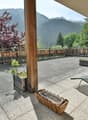 Wooden balcony or terrace with a scenic view of the mountains and forests. The balcony has a concrete floor, wooden railings, and some potted plants. There are chairs and other outdoor furniture on the balcony, suggesting it is a relaxing outdoor living s