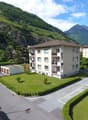 A multi-story apartment building with balconies, surrounded by well-maintained landscaping and parking spaces. The building is situated in a scenic mountainous area with lush green hills and peaks in the background.