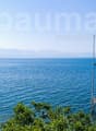 Scenic view of a calm blue sea with a sailboat in the distance, surrounded by mountains. A wooden dock or pier is visible in the foreground, with lush greenery along the shoreline.