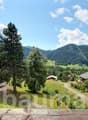 The image shows a scenic mountain landscape with lush green forests, rolling hills, and traditional alpine-style houses. The sky is blue with fluffy white clouds, creating a picturesque and serene atmosphere. The landscape appears to be in a rural, mounta