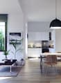 Open-concept living room and kitchen with large windows, gray sofa, wooden dining table and chairs, kitchen with white cabinets and appliances, black pendant light, and a plant decoration.