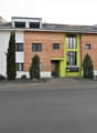 Modern apartment building with wooden panels, white walls, and green accents, featuring multiple windows and a central entrance. The building is flanked by trees and shrubs, with a paved driveway in front and several cars parked nearby.