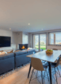 Open plan living room with L-shaped couch, fireplace, flat-screen TV, dining table, and large windows offering a mountain view.