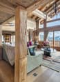Spacious open-plan living area with wooden beamed ceiling, large windows offering mountain views, plush sofas and armchairs, a fireplace, and a chandelier lighting fixture.