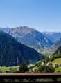 Mountain valley with lush greenery, a winding road, and distant houses, offering panoramic views of surrounding mountains.