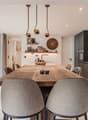 Dining room: wooden dining table, chairs, shelves with decorative items, wall clock, wooden flooring, kitchen area with sink and gas stove
