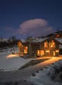 wooden house, covered in snow, stone foundation, glass windows, lit up at night, located on hillside