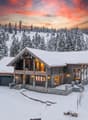 Log cabin-style house with a sloped roof, stone foundations, multiple windows, a driveway, balcony, and snow-covered surroundings