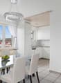 Open-concept living area with dining table and chairs, kitchen with white cabinets and appliances, large windows with curtains providing a view of the outdoors, and a modern light fixture above the dining table.
