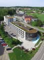 A large multi-story commercial or office building with a curved glass facade, surrounded by a parking lot and green landscaping. The building appears to be situated in a rural or suburban area with a view of a small town or village in the background.