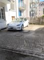 The image shows a white Tesla Model 3 electric vehicle parked on a cobblestone driveway in front of a multi-story residential building. The building has a balcony and there are trees visible in the background, indicating an urban setting.