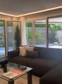 Spacious living room with large windows overlooking a private outdoor area, dark brown sofa, glass coffee table, and decorative pillows