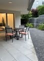 The image shows an outdoor patio area with a dining table and chairs. The patio has large sliding glass doors that lead to the interior of the home. The patio is surrounded by lush greenery, including shrubs and trees, creating a private and tranquil atmo