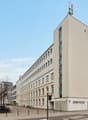 White exterior, multiple floors, rectangular windows, two entrances with text 'dreamtime', antennas on the roof, trees and bicycle stands in front, buildings behind the main building