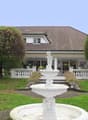 Large house with brown roof, white walls, large windows, white porch, garden with fountain, trees, and grass