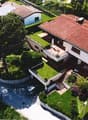 Large two-story house with red tile roof, surrounded by lush greenery and a swimming pool. The house has a balcony or terrace overlooking the landscaped garden.