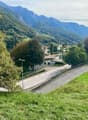 The image shows a scenic mountain landscape with a small village nestled in the valley. The scene features lush green foliage, including trees and grassy areas, as well as a winding road leading through the village. The mountains in the background are tal