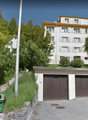 A multi-story apartment building with a garage on the ground level, surrounded by trees and greenery in a hilly, wooded area.