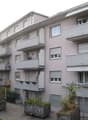 Multi-story apartment building with balconies, gray exterior, and landscaping around the building.