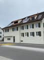 White house with solar panels on the roof, green shutters, and a brown roof