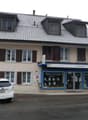2 story house, gray roof, white windows, brown shutters, Siemens store on the ground floor, white car parked in front