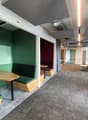 A corridor with carpet flooring, green benches, wooden tables, white walls, ceiling lights, and windows.
