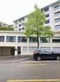 large apartment building, entrance garage with two cars parked, trees, multiple windows