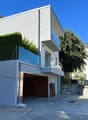 2-story modern house with a garage, balcony, and glass elements on the facade. The house has a minimalist, contemporary design with a flat roof and is surrounded by greenery.