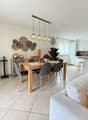 Dining room with a wooden table, gray chairs, a white couch with pillows, a black cabinet, a wine cooler, a kitchen with white cabinets, a countertop, and a hanging lamp.