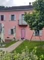 2-story pink house with shuttered windows, balcony, and a small yard with a paved walkway, garden, and outdoor furniture